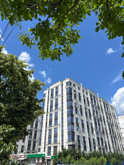 A low-angle medium shot captures a modern, white residential building in Strășeni, Moldova, under a bright blue sky dotted with white clouds. The building features a repeating pattern of dark-framed windows and some windows are tinted. Air conditioning units are visible on the exterior of the building. Trees with green foliage frame the scene from the top and left, with some of the leaves obscuring the sky. At the base of the building, there are storefronts, including one with a green awning and the "Fidesco" logo, and another with "Dulinteca" signage. A few cars are parked in front of the shops, and a single individual is walking on the sidewalk. Overhead power lines are visible against the sky on the left side of the frame.