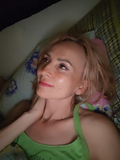 The image shows a close-up, angled shot of a blonde woman lying down, with her head resting on a floral-patterned pillow. She is looking upwards and to her left. She has light makeup on, including eyeshadow and lipstick. Her right hand is touching her neck, and she is wearing a green tank top with thin straps. The background shows a patterned blanket or comforter. The image is dimly lit. There is no visible indication of the city of Shalsky, Russia, or any urban environment.