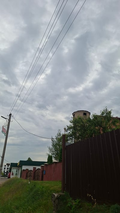 The image depicts an outdoor scene on an overcast day, likely in the city of Smolevichi, Belarus. In the foreground, tall green grass grows on a slight incline. To the right, a tall, dark brown metal fence runs horizontally across the frame. Behind the fence, a reddish-pink stucco wall with decorative metal gates is visible, bordering a grassy area. Further back, behind the wall, a building with a cylindrical brick tower topped with a darker structure is partially obscured by lush green trees. On the left side of the image, a utility pole with multiple wires stretches diagonally upwards against the cloudy sky. The wires are thin and dark. A building with a green roof and a red vehicle is visible in the far left background. The overall impression is a street-side view of residential or commercial properties under a heavy sky.