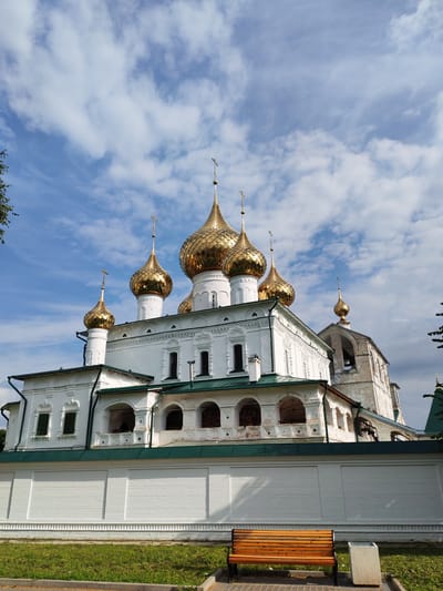 The image captures the Assumption Cathedral in Faliukovo, Russia, under a partly cloudy sky. The cathedral is a large, white building adorned with multiple golden domes, each topped with a cross. The architecture features arched windows and intricate detailing on the facade. In the foreground, a white brick wall with decorative molding runs horizontally across the scene. To the right of the wall, a wooden bench sits on a paved area, adjacent to a small concrete trash receptacle. Lush green grass covers the ground in front of the wall. The overall scene depicts a serene architectural landmark within a well-maintained outdoor space.