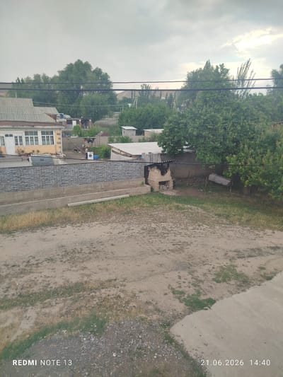 The image shows a backyard scene in Асан-Чек, Kyrgyzstan, on June 21, 2026, at 14:40. In the foreground, there is a dirt and gravel area with patches of dry grass. A concrete pathway or structure runs along the bottom right of the image. Behind this, there is a decorative stone wall with a concrete base. Further back, there is a residential building with a yellow facade, white window frames, and a gray roof. Adjacent to the building, a washing machine is visible outdoors. To the right of the building, there are several trees and a brick oven structure. The sky is overcast.