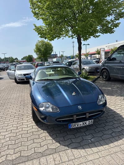 The image shows a parking lot in Ottersberg, Germany, with several parked cars. The closest car to the camera is a blue Jaguar XK with the license plate "BRV XK 18". The parking lot is paved with interlocking bricks. A tree partially obscures the view of the cars and buildings in the background. Other vehicles are visible in the row of parked cars. The sky is blue with some clouds.