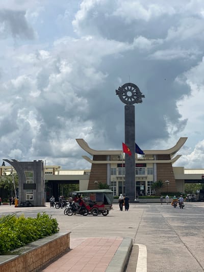 The image depicts a wide, paved area in front of a modern building complex, identified as a border crossing. The architecture of the building is distinctive with curved roof elements and prominent signage that reads "VIET NAM" in English and Vietnamese script. A tall, slender monument topped with a wheel-like emblem stands centrally in the foreground, flanked by the Vietnamese flag and another flag, possibly of Cambodia.
Several people are visible, some walking, others riding motorcycles and a three-wheeled vehicle resembling a tuk-tuk. The people are casually dressed, suggesting a warm climate. The environment appears to be a busy transportation hub, with vehicles in motion and people engaged in movement. The sky is partially overcast with dramatic clouds, indicating a dynamic weather condition. In the foreground, a low wall made of brick-like material borders a patch of green foliage, with a red-tiled walkway adjacent to it. The overall scene conveys a sense of an active international border crossing.