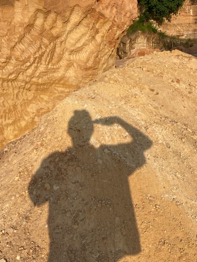 The image depicts a sandy excavation site in Ufueseni, Nigeria. The foreground is dominated by a mound of loose, light-brown soil and gravel. A distinct shadow of a person stands on this mound, their arm raised as if shielding their eyes from the sun. Behind the mound, the background reveals a layered, rocky cliff face with warm, sandy tones and visible striations. In the upper right corner, green foliage and grass grow from the cliff edge, suggesting a natural transition. The lighting indicates it is likely late afternoon, with the sun casting long shadows.