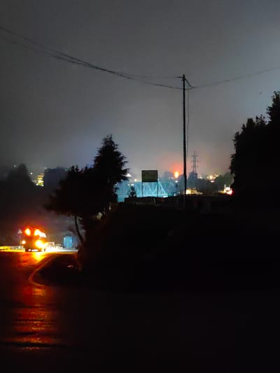 The image depicts a night scene in Kulang, India, characterized by dim lighting and fog. A vehicle with bright headlights and taillights is visible on a wet road in the foreground, reflecting the light. In the background, there are illuminated structures and what appears to be a bridge or elevated pathway. A power line stretches across the frame, supported by a utility pole. The overall atmosphere is moody and atmospheric, suggesting a potentially remote or mountainous location.