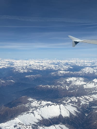 The image shows a view from an airplane window. A portion of the plane's wing is visible in the top left corner of the image. The rest of the view is of a mountain range covered with snow and the sky. The sky is blue with some white contrails. The mountain range has multiple peaks and valleys. The light is bright and the overall scene appears to be taken during the daytime.
This scene cannot be located in the city of Fischamend, Austria, because Fischamend is a town located on a plain. It does not have mountainous terrain.