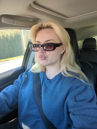 A fair-skinned individual with long, wavy blonde hair and dark, rectangular sunglasses is photographed seated in the driver's seat of a vehicle in Area D (Elaho/Garibaldi), Canada. The individual wears a blue long-sleeved sweater and a black seatbelt. A small sun-like tattoo with radiating lines is visible on the upper left chest. A dangle earring is present in the left ear. The individual's lips are pursed in a pouting expression. The vehicle interior shows a light-colored headliner and a sunroof. Outside the driver's window, green foliage and a utility pole are visible under daylight conditions.