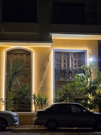 This image depicts a nighttime scene on a street in Al Hadaba, Egypt. The primary subjects are the exterior of a building with two windows, a dark-colored car parked on the right side of the frame, and some greenery in front of the building. The building's facade is a warm, sandy yellow, accented by modern LED strip lighting that outlines the windows and a curved architectural feature. The windows themselves appear to be made of dark wood with a decorative metal grate in the upper window.
The upper window is larger and rectangular, while the lower window is smaller and arched. Plants, including palm fronds and leafy branches, are situated in front of and around the windows, suggesting a courtyard or garden area. The dark-colored car is partially visible, angled towards the building. Its headlights are off, and its presence indicates that the scene is likely in a residential or commercial area.
The time of day is clearly night, as evidenced by the dark sky and artificial lighting. The lighting on the building creates a warm and inviting ambiance. There is no visible text in the image. The overall impression is one of a peaceful, well-lit evening in a town or city.