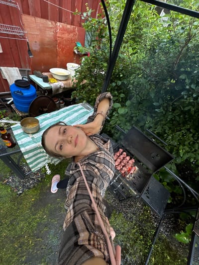 An overhead shot captures a woman posing for a selfie in an outdoor setting, likely a backyard, in a rural area or the outskirts of the city of Leningradskaya, Russia. The woman, wearing a plaid shirt and pink clogs, is lying on a green and white striped blanket. To her left, a metal pot with food and a bottle of beer are visible. In the background, a black metal grill with skewers of raw meat is in operation, emitting smoke. The surroundings consist of lush green foliage and a rustic wooden wall, suggesting a casual, domestic atmosphere. The ground appears wet, indicating recent rainfall.