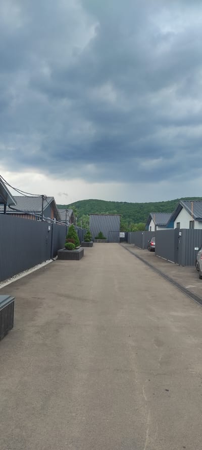 The image shows a row of modern, dark gray buildings with dark roofs, likely apartments or townhouses, situated on a hillside in Tulskiy, Russia. A paved driveway runs alongside the buildings. The buildings are in a linear arrangement, with some featuring white accents. Green trees cover the hillside behind the buildings. The sky is overcast with heavy, dark gray clouds, suggesting an impending storm or recent rain. There are several planters with small evergreen trees placed along the driveway. One of the buildings has a car parked in front of it. The overall scene conveys a sense of modern, somewhat minimalist architecture set against a natural, hilly landscape. There is no visible text in the image. The time of day appears to be daytime, but the weather conditions make it feel gloomy.