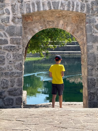 The image depicts a young boy standing in front of an arched stone gateway, looking out at a body of water. The gateway is constructed from rough-hewn stones, with a slightly pointed arch. Through the arch, a lush green tree with some small red fruits is visible. Beyond the tree, the water reflects the greenery and the sky. On the far side of the water, a set of stone stairs ascends. The boy is wearing a bright yellow t-shirt, dark shorts, and a baseball cap. His stance suggests he is observing something in the water, possibly a duck which is faintly visible. The ground in the foreground is paved with irregular stone slabs. This scene is likely located at the source of the Trebišnjica River in Trebinje, Bosnia and Herzegovina, known for its picturesque stone architecture and natural beauty.
