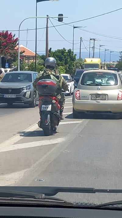 A man wearing military camouflage and a helmet rides a motorcycle through a street in Marmaroto, Greece. He is surrounded by other vehicles, including a car and a yellow bus. The man is carrying a large backpack on his back. The sun is shining, suggesting it is daytime. The road appears to be paved and marked with white lines. In the background, there are trees and buildings, with a body of water visible in the distance. The license plate on the motorcycle reads "PZI 5994". The license plate on the car in front of him reads "MHL 8080". The license plate on the silver car in the foreground reads "AIX 2215".