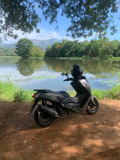 In Buduruwagala, Sri Lanka, a grey scooter is parked on a dirt path next to a body of water. The scooter is positioned in the foreground, angled slightly towards the left. A black helmet is placed on the handlebars. The water's surface reflects the surrounding lush greenery and distant hills under a clear blue sky with scattered clouds. Lily pads and aquatic plants are visible on the water. The environment is natural and serene, with trees and foliage lining the banks of the water.