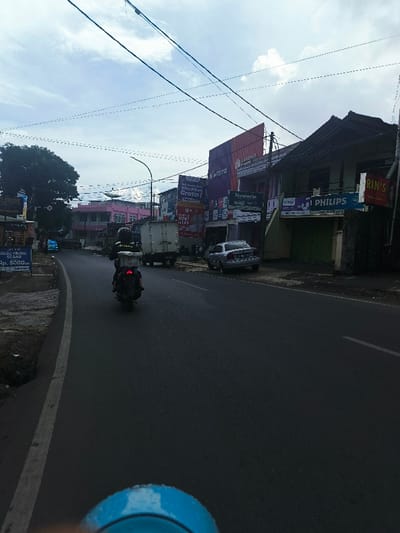 The image shows a street in Lembang, Indonesia. The coordinates, -6.816 latitude and 107.619 longitude, indicate a location in a residential area. The scene is dominated by a two-lane road with a white dividing line. A motorcycle with a rider is visible, moving away from the camera on the right lane. The street is lined with shops on the right side, with signs for businesses like "Kivero," "Hanroaches," "Philips," and "Rin's Laundry." The buildings have a light gray color and appear to be made of brick or concrete. There are several power lines stretching across the sky above the street. On the left side of the road, there are more buildings, a few cars, and a small sign for a store that sells vegetables. The overall scene is one of typical Indonesian suburban life. The sky is a light blue with some clouds. The weather appears to be overcast.