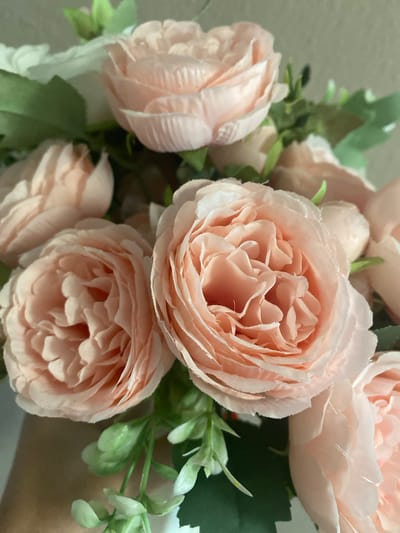 This is a close-up, eye-level shot of a bouquet of artificial, pale pink roses with green foliage. The flowers have many delicate, layered petals. The background is a soft, neutral beige wall. The lighting is even and bright, suggesting it was taken indoors during the day. There is a hand visible on the right side of the image, gently holding the bouquet, suggesting someone is presenting or arranging the flowers. The overall mood is soft, romantic, and delicate.