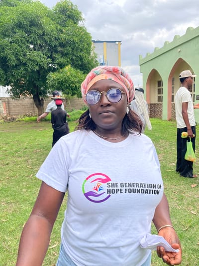 A group of people are gathered outdoors on a cloudy day in Unguwar Shanu, Nigeria. The main subject is a woman in the foreground, wearing a white t-shirt with the text "SHE GENERATION OF HOPE FOUNDATION" and a logo. She has a headscarf on and is wearing sunglasses. She is holding a crumpled piece of paper in her right hand.
In the background, a person wearing a white t-shirt and black pants is holding a bright yellow bag. Further in the background, a building with a light green facade and arched windows is visible, suggesting a public or institutional setting. There are also trees and grassy areas, indicating an outdoor environment. The overall mood appears casual, possibly related to a community event or outreach program organized by the foundation mentioned on the t-shirt.