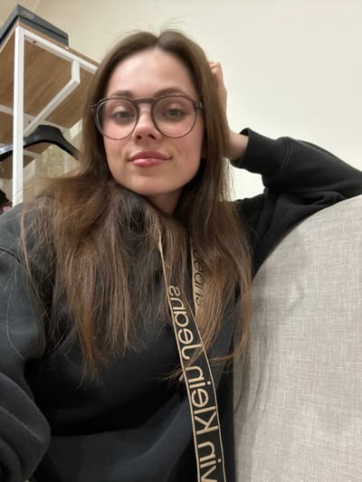 A female subject is seated indoors in what appears to be a residential setting in Scafati, Italy. She has long, straight brown hair and wears round, dark-framed glasses. She is dressed in a dark-colored hooded sweatshirt, with a beige strap displaying the inverted text "calvin klein jeans" traversing her chest. Her right hand is placed at the back of her head, while her left arm rests on a light grey, textured upholstered surface. The subject is looking directly at the camera with a faint smile. In the background to the left, a white metal shelving unit is visible, holding a dark rectangular object on its top shelf and a black clothes hanger on a lower support. The wall in the background is a plain light color.