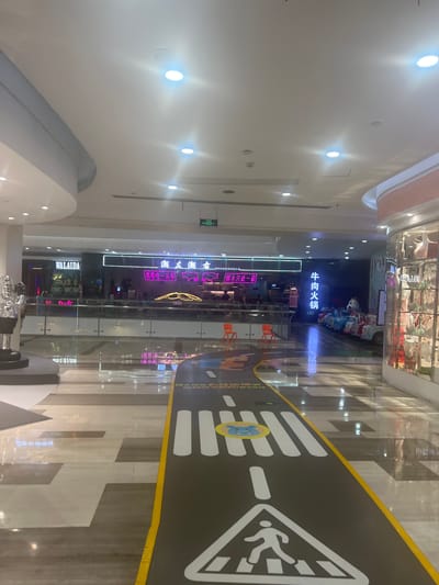 The image shows the interior of a shopping mall in Liandu District, China. In the foreground, a children's play area is designed to resemble a road with a crosswalk. The road features white painted lines for lanes and a pedestrian crossing symbol with a cartoon animal. In the middle ground, there is a row of colorful children's ride-on cars. Beyond the play area, a glass partition separates the walkway from a dining establishment with neon signage in Chinese characters, including "牛肉火锅" (beef hot pot). To the left, a store with the "WALAIDA" logo is visible. The mall environment is illuminated by overhead lights and reflects off the polished floor. In the background, more shops and dining areas can be seen.