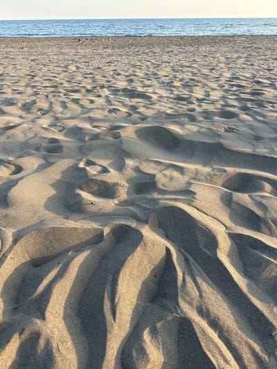 The image depicts a sandy beach in Donji Štoj, Montenegro. The foreground is dominated by sand, marked with footprints and wave-like patterns created by wind or water. The sand's texture is detailed, with shadows accentuating the ridges and depressions. In the background, a strip of water, likely the Adriatic Sea, is visible, bordered by a light-colored sky, suggesting either dawn or dusk. There are no people or objects other than sand and water in the frame. The scene captures a natural, unpopulated coastal environment.