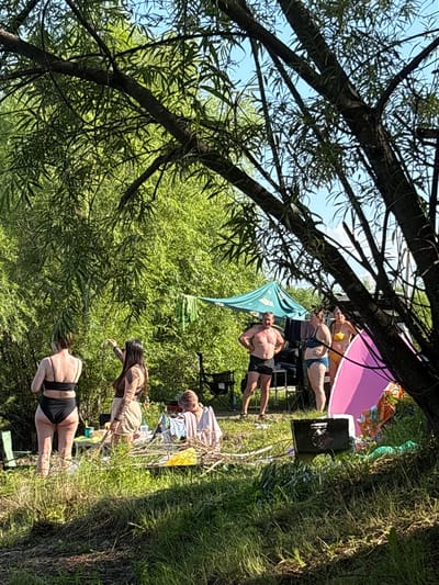 The image depicts a group of people gathered outdoors in a grassy area with dense green foliage and trees. In the foreground, a large tree trunk and branches obscure part of the view. Several individuals are visible in the mid-ground, some wearing swimwear. There is a green tent or canopy set up, and various items are scattered on the ground, including what appears to be a picnic table with some objects on it, a grill, and folded fabric. The overall scene suggests a casual outdoor gathering or picnic.