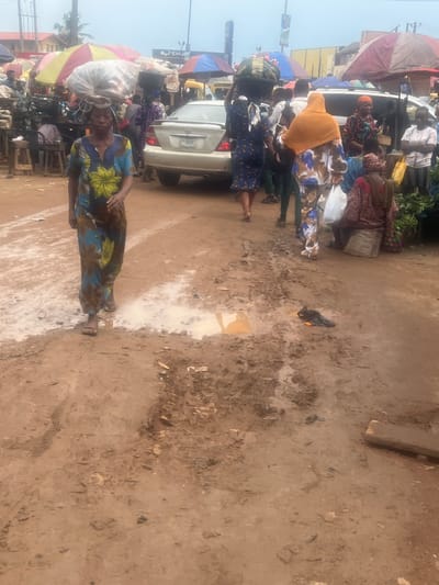 The image depicts a bustling street scene in Egan, Nigeria. A woman in a patterned blue and yellow dress carries a large white load on her head, walking on a muddy, uneven ground. A white car is parked nearby, with people gathered around it. Several individuals carry items on their heads, and colorful umbrellas provide shade from the sun. The environment appears to be a marketplace, with various vendors and pedestrian traffic. The overall scene conveys a sense of daily life and commerce in a crowded urban environment.