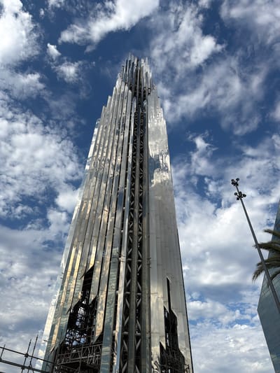 A tall, modern skyscraper with a faceted, metallic exterior stands against a cloudy blue sky. The building's surface is highly reflective, mirroring the sky and clouds. The structure appears to be under construction, with scaffolding visible at its base and a visible metal framework within some of the building's sections. To the right of the skyscraper, a tall lamppost with multiple lights stands, and a portion of another glass-faced building and a palm tree are visible. The image is taken from a low angle, emphasizing the height of the skyscraper. The location is identified as Garden Grove, United States of America.