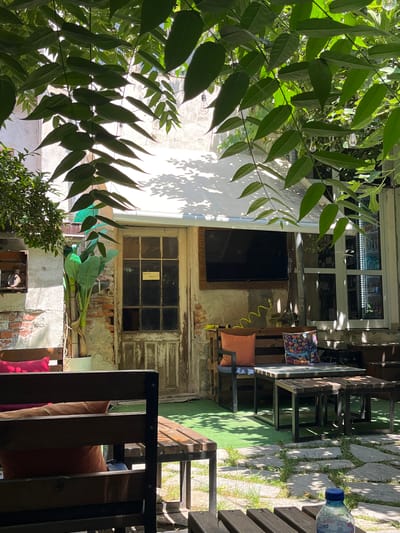 The image depicts an outdoor seating area with a rustic and eclectic ambiance, likely a cafe or restaurant. The scene is dominated by lush green foliage in the foreground, framing a view of the seating area and the building behind it. A white wall with shadows cast by the leaves is visible on the left.
In the center, a weathered building features a large window with multiple panes, some of which appear to be broken or in disrepair, adding to the rustic charm. Below this window, a dark screen is mounted on the wall, and a yellow coiled hose hangs nearby. To the right of the building, a wooden pallet-style bench is adorned with two cushions, one patterned and the other orange. Further to the right, a wooden table and chairs are arranged on a surface that appears to be a combination of flagstones and artificial turf. A partially visible bottle of water with a blue cap rests on one of the wooden benches.
The overall impression is one of a relaxed, bohemian outdoor space. The dappled sunlight suggests it is daytime, and the greenery implies pleasant weather. The setting in Gotse Delchev, Bulgaria, contributes to a sense of place, suggesting a Mediterranean or Balkan influence in the design and atmosphere. The image captures a quiet moment, with no people actively engaged in activities, but the presence of seating and a table implies a space for social gathering. A small label with handwritten text is visible on the window pane.