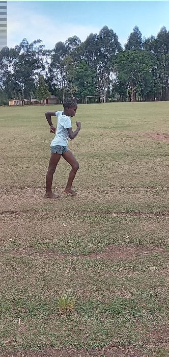 The image shows a young person, likely a teenage girl, in a white shirt and denim shorts, dancing in a grassy field in Bungoma, Kenya. The girl has dark skin and her hair is braided and tied with a white string. She is dancing on the grass with her arms outstretched and her feet moving in a rhythmical manner. Her face is obscured by the sun's glare. The location, based on the coordinates provided, is a field near a group of pine trees. The trees are tall and slender, their foliage casting long shadows on the ground. The field is covered in a mix of green and brown grass, and there are no other people or objects visible in the image.