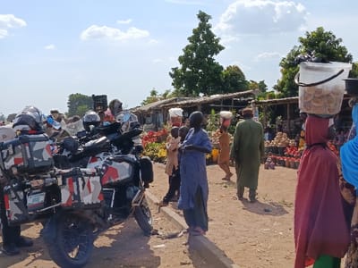 The image depicts an outdoor market scene in Jere North, Nigeria, under a bright, partly cloudy sky. The ground throughout the scene is unpaved dirt.
In the foreground left, a heavily loaded adventure touring motorcycle with camouflage-patterned panniers is parked. Multiple motorcycle helmets are visible on or around the bike, and its rear license plate reads "913G" from "KADUNA." A person's lower legs, clad in dark trousers, are visible near the motorcycle's rear wheel. A low, concrete curb runs horizontally across the foreground right.
The midground features several individuals walking or standing. A person in a light blue tunic stands near the curb, facing left. Further back, another person in a light tan garment walks left, balancing a white basin on their head. An individual in a dark olive-green robe walks away from the camera. Towards the right, a person wearing a red full-body garment carries a large, transparent plastic bucket filled with light-colored, round food items on their head; another person in blue attire is partially visible beside them. Children are visible among the market-goers.
The background is dominated by multiple market stalls constructed from wood and sheet materials. These stalls display abundant fresh produce, prominently featuring large quantities of bright red tomatoes arranged in buckets and piles on the ground. Green trees are visible beyond the stalls, under the sky.