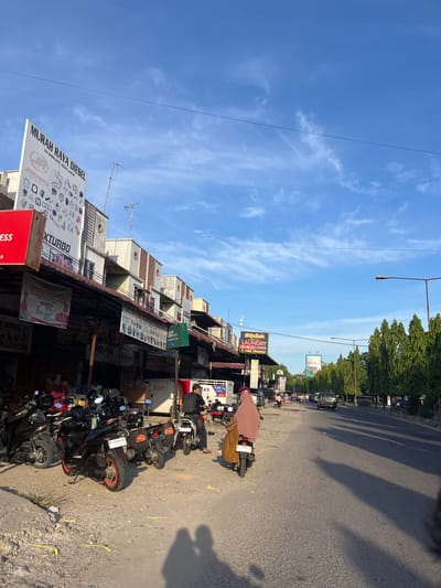 The image shows a street scene in Langsa, Indonesia. In the foreground, several motorcycles are parked on a gravelly shoulder. A person wearing a brown headscarf and a yellow dress is riding a motorcycle down the street. Further down the street, there are buildings with signs advertising various businesses, including "MURAH RAYA DIESEL", "XTURBO", and "LAUNDRY EXPRESS". There are trees lining the opposite side of the street, and the sky is blue with scattered clouds. The sun is low in the sky, casting long shadows across the road.