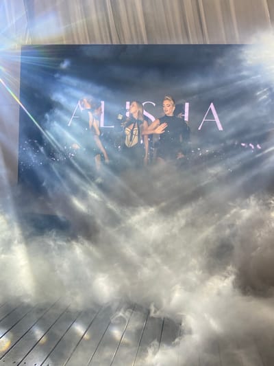 The image captures a stage performance with three women in the foreground, silhouetted against a dark backdrop with the word "ALISIA" in large, pink, illuminated letters. The stage is filled with atmospheric smoke and dramatic lighting, with visible beams of light cutting through the haze. The women appear to be singers or performers, dressed in dark, stylish outfits. One woman in the center is wearing a black, sequined top and shorts, with her hand raised. To her left, a woman is in a strapless, cream-colored corset with black straps, and to her right, another woman in a dark outfit is partially obscured. The stage floor, visible at the bottom of the image, appears to be made of wooden planks and is also covered in smoke. The overall impression is one of a vibrant and energetic performance. The location in Калодзішчанскі сельскі Савет, Belarus, is not discernible from the image itself.