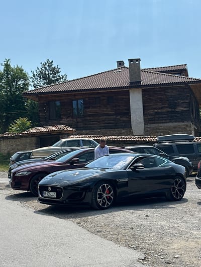 In Arbanassi, Bulgaria, a black Jaguar F-Type coupe is parked on a gravel lot in front of a traditional wooden house with a tiled roof. The car has a license plate reading "B 935 JAG." To the left of the Jaguar, a maroon Jaguar F-Type is visible. Behind these cars, a vintage gold sedan and a dark-colored SUV with a roof box are parked. A man in a white t-shirt stands between the two Jaguars, looking down. The scene is set under a clear blue sky, with trees and part of a stone wall visible in the background.