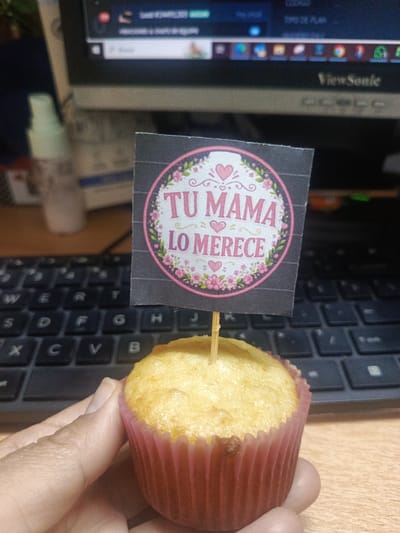 A hand holds a cupcake with a pink paper liner and a toothpick in the center holding a square black paper sign. The sign has a floral wreath design with the words "TU MAMA LO MERECE" written in the center. In the background, there is a black computer keyboard, a monitor with "ViewSonic" visible, and a bottle of hand sanitizer. The overall setting appears to be indoors, possibly an office or workspace. The location in Guayaquil, Ecuador is not evident from the image.