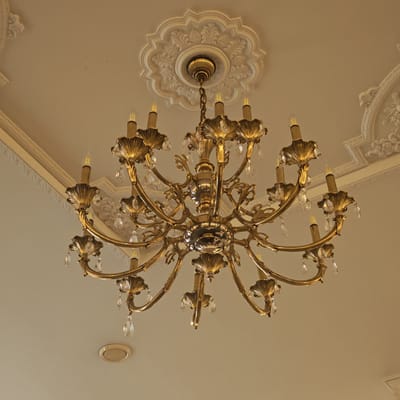 An ornate, multi-armed chandelier dominates the center of the image, viewed from below against a light-colored ceiling. The chandelier is crafted from polished brass or a similar gold-toned metal, featuring numerous gracefully curved arms, each supporting a faux candle-style light bulb and adorned with intricate, leaf-like metallic details. Clear, faceted crystal droplets hang from many points, adding sparkle and reflection.
The chandelier is suspended from an elaborate white plaster ceiling medallion, which features a detailed, lobed design with botanical or classical scroll motifs. Surrounding the central medallion, and extending towards the corners of the ceiling, are additional decorative plaster moldings. These moldings exhibit complex patterns, suggesting a classical or baroque architectural style. The ceiling itself is a plain, light, neutral color, providing a subtle backdrop for the elaborate fixtures.
The scene depicts an interior space, likely a formal room, characterized by its traditional and elegant decor. A small, circular, flush-mounted fixture, possibly another light or a ventilation unit, is visible on the ceiling towards the bottom of the frame. There are no people or specific activities taking place, and no visible text. The overall impression is one of refined grandeur, fitting for a historic or well-appointed building in a location like Ново-Никольское, Russia.