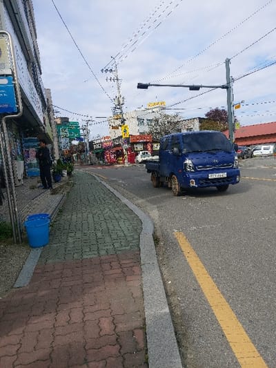 This image depicts a daytime street scene in 강화군, South Korea, under an overcast sky. The foreground shows a paved sidewalk, composed of reddish-brown and green interlocking pavers, bordered by a grey granite curb. A blue plastic bucket rests on the sidewalk adjacent to a commercial building facade on the left. A man in dark clothing stands near the building.
The midground features an asphalt road with a solid yellow line marking its edge. A blue Kia Bongo 3 flatbed truck, license plate 80가 8547, is captured in motion on the road, moving away from the viewer. Above the road, a dark traffic light is mounted on a grey pole, and a utility pole festooned with transformers and numerous overhead power lines rises prominently.
In the background, various multi-story buildings line the street, featuring numerous storefronts with Korean signage, indicating a commercial district. Other vehicles are visible in the distance. The sky above is largely grey with brighter patches.