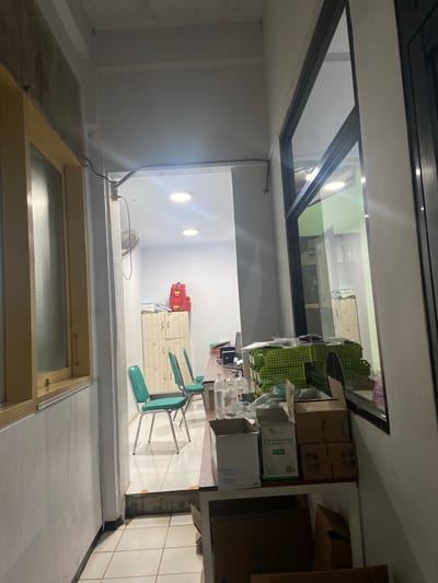 The image depicts an indoor scene, likely a medical clinic or office, in Margoyoso, Indonesia. A narrow hallway leads to a well-lit room with multiple cabinets, chairs, and a desk. In the foreground of the hallway, a window with a frosted glass pane runs along the left wall. The hallway floor is tiled, and a faint orange mark is visible on the tiled step leading into the main room.
The main room is visible through an opening. Two teal chairs with metal frames are positioned in front of a light-colored cabinet with several drawers and a desk. Two red medical bags are placed on top of the cabinet. A ceiling fan is mounted on the wall in the hallway, and circular fluorescent lights illuminate the room.
On the right side of the image, a large glass window reflects the interior of the room and the exterior. Stacks of green plastic baskets are piled on a shelf in front of the window, along with several cardboard boxes and bottles. One of the boxes is labeled "GP Care 3-Way Stopcock with Extension Tube 50 Pcs". The overall impression is of a functional, albeit somewhat cluttered, workspace.