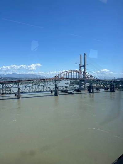 The image depicts the Alex Fraser Bridge and the New Westminster Bridge in New Westminster, Canada, spanning the Fraser River. The sky is a clear blue with scattered white clouds. The water of the Fraser River is a murky, light brown color, reflecting the sky. Several cargo ships are visible in the river. The Alex Fraser Bridge, a cable-stayed bridge, is in the background with its tall, grey towers. In the foreground is the older, truss-style New Westminster Bridge, painted in sections of green and orange. The bridge structure is complex, with multiple levels and supports. On the far left, a cityscape with mountains in the background is visible. The image appears to have been taken from a vantage point above the river, possibly from another bridge or a tall building, with reflections of the sky visible on a surface in the foreground.