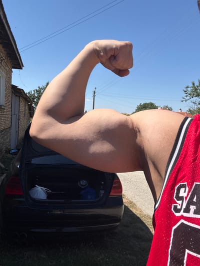In Konop, Bulgaria, a person in a red mesh jersey flexes their bicep while standing outdoors. A black car with an open trunk is parked behind them, and a brick building is visible to the left. The sky is clear blue, and the sun is shining brightly.