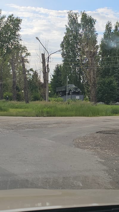 A monument featuring a military truck is displayed in a grassy area in Kamensk-Uralsky, Russia. The truck, likely a ZIL-157, is green and mounted on a pedestal. It is surrounded by trees and a blue sky with clouds. A street lamp is visible to the left of the truck. The foreground shows a paved road.