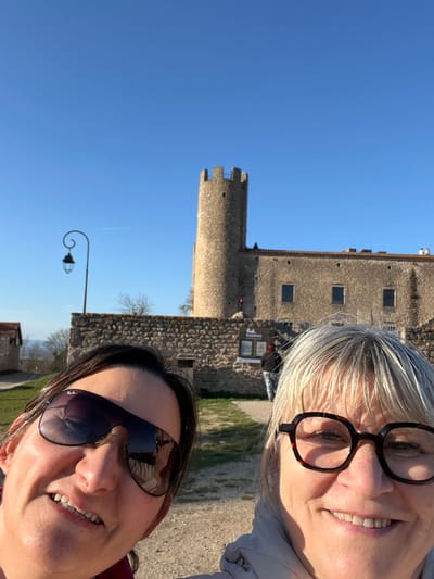 A selfie photograph captures two adult women in the foreground against the backdrop of the Château de Chambles in Chambles, France, under a clear blue sky.
The woman on the left has dark hair, wears dark Ray-Ban sunglasses, and is smiling towards the camera. She is visible from her forehead to her upper chest. The woman on the right has blonde hair, wears tortoiseshell-framed eyeglasses, and is also smiling. She is visible from her forehead to her upper chest.
In the mid-ground, a dirt path leads towards a stone wall. A black decorative street lamp with a scrolled arm and lantern is positioned to the left. A small, light-colored building with a dark roof is partially visible further left. On the stone wall, a sign with dark text on a light background reads "Le Essario." A third individual in dark clothing is partially visible standing near the base of the castle wall to the right of the tower.
In the background, the Château de Chambles stands prominently. It features a tall, round stone tower with crenellated battlements on the left. Adjacent to the tower is a longer, lower stone building with several rectangular windows. The structures are built from light-colored stone. Sparse, bare trees are visible behind the wall and to the left of the castle. A distant landscape is visible on the far left horizon. The sky is bright blue with a faint white contrail visible.