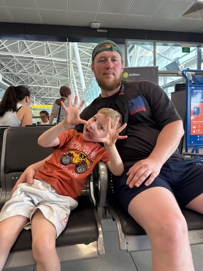 The image shows a man and a young boy sitting in an airport terminal. The man is wearing a dark t-shirt and a baseball cap, and has a beard. The boy is wearing an orange t-shirt with a graphic of a monster truck and the word "EXTREME," and light grey shorts. He is sticking his tongue out and making a funny face. The background shows airport seating, a large window with a view of the sky and building structures, and a sign for S7 Airlines. The scene appears to be within the Bol'shie Kabany airport, Russia.