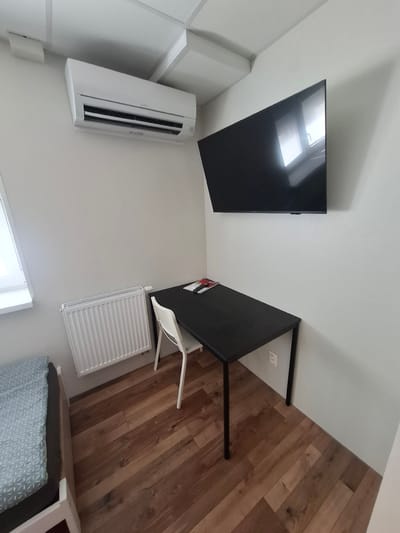 The image depicts a sparsely furnished room, likely a small apartment or dorm room, in Bratislava, Slovakia. On the wall, a modern white air conditioning unit is mounted near the ceiling, and a large, black flat-screen television is also wall-mounted above a simple black desk. A white chair is positioned at the desk. To the left of the desk, a white radiator is installed against the wall, and next to it is a window with blinds. In the foreground, a portion of a bed with a patterned blue and grey duvet is visible. The room has wooden plank flooring and white walls. There are no people in the image.