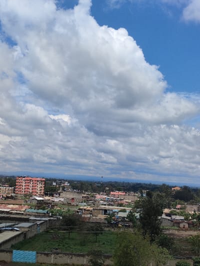 This image presents an elevated, panoramic view of a densely developed urban landscape within Juja ward, Kenya, under a dynamic sky dominated by large cumulus clouds and patches of clear blue.

The foreground features a concrete perimeter wall, behind which lies an unkempt green plot of land with sparse vegetation. To the immediate left of this plot, a light blue corrugated metal fence is visible. The midground and background are densely populated with structures. A prominent multi-story building with reddish-orange exterior walls and multiple balconies stands on the far left, surrounded by numerous single to two-story buildings, many featuring corrugated metal roofs of varying conditions, interspersed with green trees and bushes. Towards the right, an incomplete or grey-walled multi-story structure is observable. The overall residential and commercial architecture appears varied, indicative of ongoing development and established settlements. In the distant background, a slender communication mast or tower is visible rising above the tree line, with faint outlines of hills or mountains extending across the horizon. No specific individuals or discernible actions are visible within the frame.