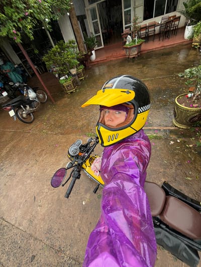 The image shows a person wearing a purple rain jacket and a black and yellow helmet, sitting on a yellow motorcycle. The person is holding the camera, taking a selfie. The motorcycle is parked on a wet, concrete surface, which appears to be an outdoor area. In the background, there are several potted plants, some motorcycles, and buildings that suggest an urban or semi-urban environment. The presence of rain is evident from the wet ground and the falling raindrops. This scene is located in Hướng Phùng Commune, Vietnam, likely in front of a building that may be a shop or residence, given the presence of outdoor seating and parked vehicles.