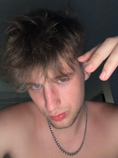 A close-up, low-angle, selfie-style portrait of a young Caucasian male is presented. The subject, positioned center-frame, has light brown, tousled hair, pale skin, and light blue eyes. A small red blemish is visible on his nose. He is wearing a silver chain necklace. His right hand is raised, with fingers touching his ear. The background is dark and out of focus, with hints of a dark wall and a faint vertical line suggesting a doorway or window frame. The overall lighting is dim, with the subject's face illuminated from the front. There are no identifiable landmarks or features indicating a specific location within Vanino, Russia.