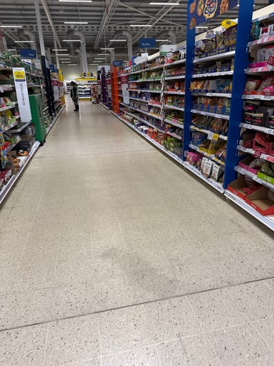 The image shows an aisle in a Tesco supermarket in Hull, United Kingdom. The shelves are stocked with various food products, including pasta, rice, and other items labeled "All World Foods". Price tags are visible on the shelves. A person wearing dark clothing and a fluorescent vest is seen in the distance down the aisle. The floor is a light-colored tile. The ceiling has visible support beams and lighting fixtures.