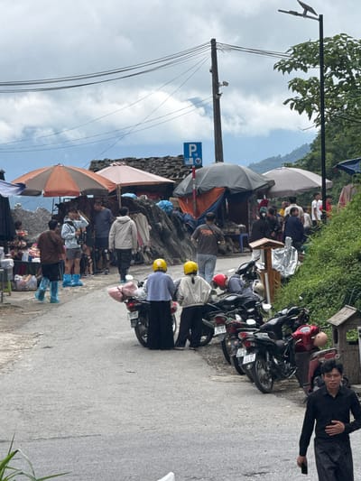 A ground-level, medium-shot image captures a street scene in Yên Minh Commune, Vietnam. The viewpoint is along a roadside, where people are engaged in street commerce under several umbrellas, and motorcycles are parked. The photograph is taken at an angle, with the foreground slightly higher than the background.

On the right, a man is lying on the ground beside the parked motorcycles, holding a cell phone in his right hand. Behind him are several motorcycles, their license plates partially visible.  Several people are near the vehicles. The license plates have Vietnamese writing.

On the left side of the scene, under the shade of a large orange umbrella, people are observed shopping at a roadside market. Many of them are wearing blue plastic shoe covers, indicating potential muddy conditions. Further in the distance, more people are congregated around tables and vendors. An old parking sign, “P 18,” is attached to the side of a stone wall. Overhead, power lines stretch across the sky, running diagonally from the left side of the scene toward the background.

The environment appears to be a mountainous region, as evidenced by the rising terrain and lush greenery in the background. The sky is filled with gray clouds, suggesting overcast weather.
