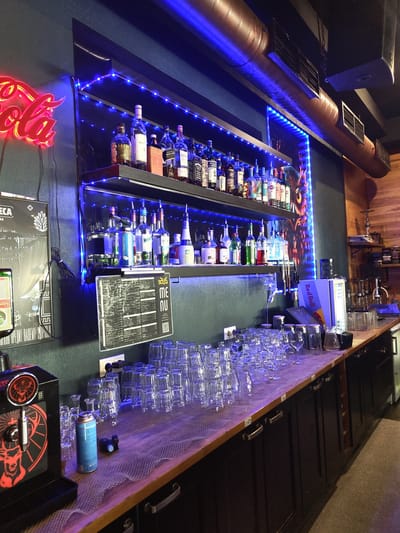 This image shows a bar interior, likely located in Zhukovsky, Russia, given the text on the menu board. The bar is dimly lit, with prominent blue LED lighting illuminating shelves of liquor bottles and rows of glassware. A neon red Coca-Cola sign is visible on the left wall. The bar counter is covered with a white mesh-like material and features stacks of clean drinking glasses, a beverage dispenser labeled "Jägermeister", and a blue CO2 canister. Behind the counter, shelves are stocked with various alcoholic beverages, including bottles of whiskey, vodka, and liqueurs. A large menu board is displayed on the wall. The overall atmosphere suggests a modern and well-equipped bar or lounge.