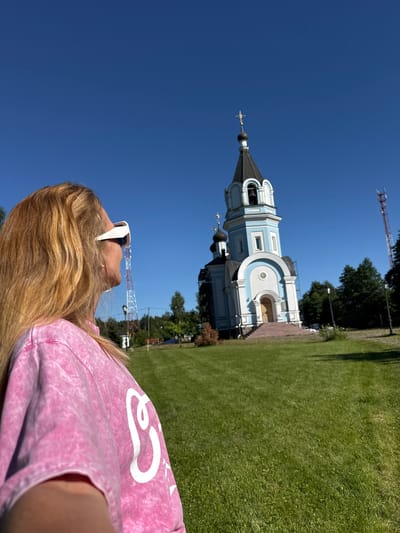 An outdoor, eye-level, medium shot shows a blonde woman from the shoulders up, facing right and looking toward a blue and white domed church in the background. The woman is wearing a pink tie-dye t-shirt and white-rimmed sunglasses. Her blonde hair is shoulder length. The church, in the middle ground, has a tall bell tower topped with a gold cross and a black, onion-shaped dome. The church’s main dome is black and also topped with a gold cross. A red and white cell tower is visible to the left of the church, and a dense line of trees forms the background. The scene is set on a bright, sunny day with a clear blue sky, and the foreground is a grassy lawn.