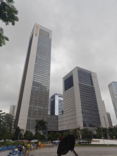 The image shows a street-level view of modern skyscrapers in the Shunde District of China. The sky is overcast. In the foreground, a paved area is visible with a black parabolic-shaped object mounted on a pole in the center. To the left, rows of blue bicycles are parked. Further back, there is a street with several cars, including red and white vehicles. Trees and green spaces are interspersed between the buildings.

The prominent buildings include a tall, slender skyscraper with golden Chinese characters on its upper section, and a wider, rectangular skyscraper to its right, featuring the "InterContinental" logo. Another glass-fronted building is visible between these two. In the distance, other high-rise buildings can be seen. A large white structure with golden Chinese characters, possibly a landmark or sign, is situated in the mid-ground on the right side of the image. People are visible near the parked bicycles and on the street, some appearing to be walking or standing. The overall impression is of a bustling urban environment.